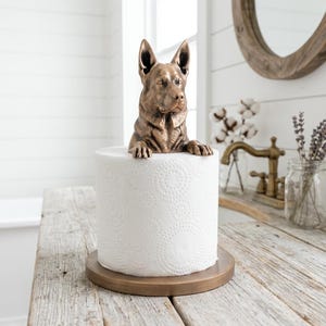 May include: A bronze-colored German Shepherd dog figurine perched atop a white toilet paper roll. The dog sculpture features detailed fur texture, pointed ears, and front paws resting on the top edge of the roll, which sits on a circular base.