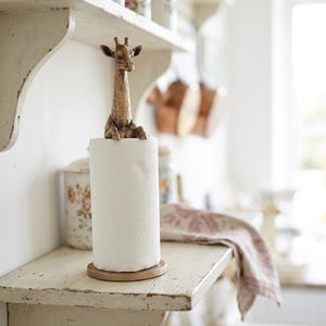 May include: A vertical paper towel holder featuring a bronze-toned metal giraffe figurine topper. The giraffe has a textured neck and sits atop a cylindrical roll of white paper towels, which rests on a circular base made of matching bronze-toned metal.