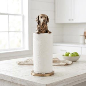 May include: A decorative paper towel holder featuring a bronze-coloured dog sculpture. The dog, a Golden Retriever, sits atop a white paper towel roll. The holder has a bronze base. The kitchen setting includes a white countertop and a window. The paper towel roll is approximately 25 cm tall.
