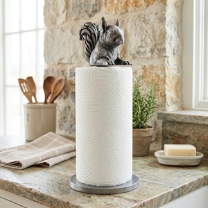 May include: A metal paper towel holder featuring a dark grey, sculpted squirrel figure perched atop a cylindrical post. The base is circular and flat, supporting a standard roll of white paper towels with a textured, embossed pattern.