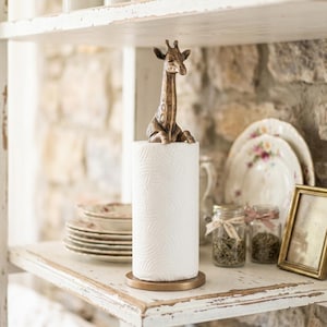 May include: A bronze-toned metal paper towel holder featuring a sculpted giraffe figure at the top. The holder has a circular base and a vertical rod that supports a standard roll of white paper towels with a textured, embossed geometric pattern.