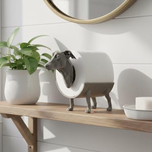May include: A novelty toilet roll holder in the shape of a greyhound dog. The dog's head and neck emerge from the centre of the roll. The holder is grey and brown and sits on a wooden shelf. A plant in a white pot is to the left.