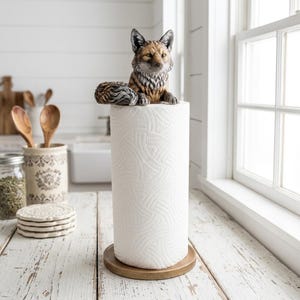 May include: A paper towel holder featuring a sculpted fox figure with brown, grey, and white fur tones resting on top of a white paper towel roll. The base is a circular wooden disc that supports the roll, providing a stable foundation for the kitchen item.