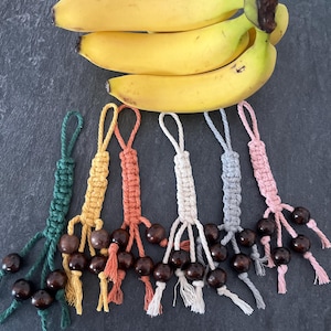 May include: A cluster of ripe yellow bananas sits above six handmade macrame keychains. Each keychain is a different colour: green, yellow, orange, white, grey, and pink. Dark brown wooden beads and tassels decorate each keychain, creating a natural and earthy aesthetic.