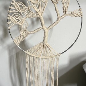 May include: A cream-coloured macrame wall hanging featuring a tree design constructed from knotted cord. The tree is centred within a circular silver-toned metal hoop, with a series of long, loose vertical cords extending from the base of the trunk.