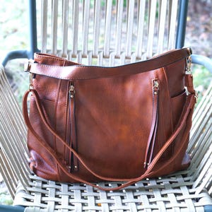 Puede incluir: Un bolso de hombro marrón de cuero sintético con múltiples bolsillos y una correa larga ajustable. El bolso tiene dos compartimentos con cremallera con tiradores decorativos y está sobre una silla tejida.