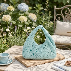 May include: A light blue crochet handbag with a floral pattern sits on a wooden board. The bag has a curved handle and a soft, textured appearance. A matching teacup and saucer are on the table, along with a pair of sunglasses and a book.