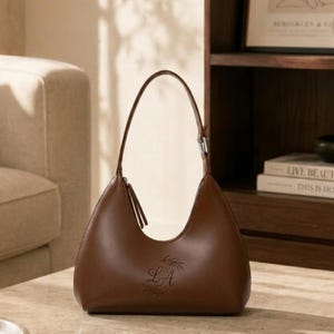 May include: A brown leather shoulder bag with a curved design. The bag has a single handle and a zipper closure. The initials "LA" are embossed on the front, along with a decorative leaf design. The bag is sitting on a marble surface.