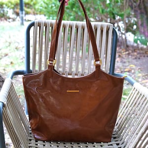 May include: A brown leather tote bag with long handles and gold-colored hardware. The bag has a glossy finish and a rectangular shape. The bag is sitting on a chair with a light-colored woven seat and back.