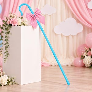May include: A bright blue shepherd's crook featuring a large, satin-finish pink bow tied at the top. The crook is positioned leaning against a white rectangular pedestal, creating a clear view of the smooth, curved handle and long shaft.