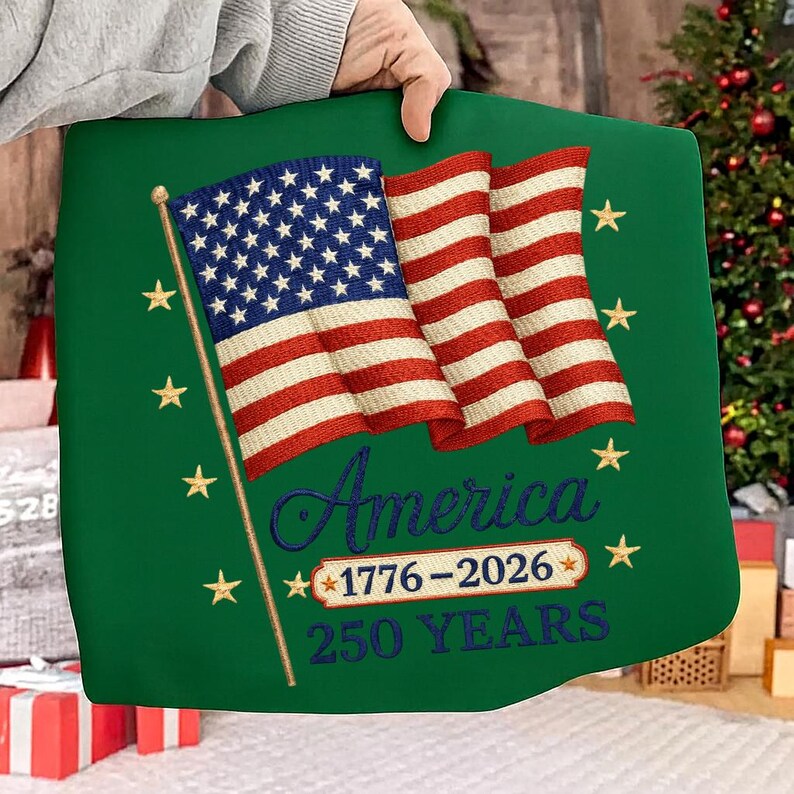 Puede incluir: Un coj&iacute;n verde con un dise&ntilde;o bordado de la bandera estadounidense. La bandera tiene rayas rojas y blancas y estrellas azules. La palabra "America" est&aacute; bordada en cursiva, con "1776-2026" y "250 YEARS" debajo. Estrellas doradas est&aacute;n bordadas alrededor de la bandera.