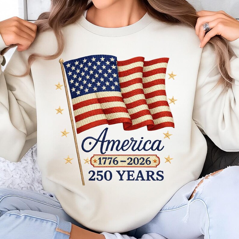 Puede incluir: Sudadera color crema con una bandera estadounidense estilizada con rayas rojas y blancas y un campo de estrellas azul. La palabra "America" est&aacute; escrita en cursiva debajo de la bandera, con "1776-2026" y "250 YEARS" en una pancarta debajo.