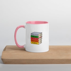May include: White ceramic mug with a pink interior and handle. The mug features a stack of books with titles like "Enemies to Lovers" and "Prophecy". The mug is placed on a wooden surface.