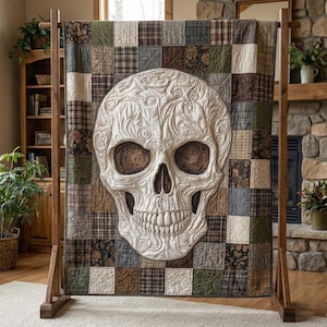 Op de afbeelding: Een grote, decoratieve quilt met een gedetailleerd schedelontwerp. De schedel is crèmekleurig met ingewikkelde reliëfpatronen en bruine oogkassen. De achtergrond van de quilt is een patchwork van vierkanten in diverse aardetinten.