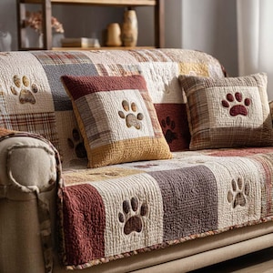 Puede incluir: Una funda de sofá patchwork y cojines a juego en tonos beige, marrón y burdeos. La funda y los cojines presentan huellas de patas bordadas. La funda del sofá está colocada sobre un sofá beige.