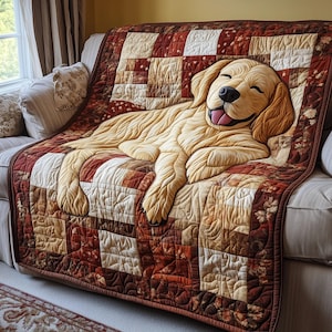 Op de afbeelding: Een handgemaakte quilt met een ontwerp van een lachende golden retriever. De quilt is gemaakt van verschillende vierkanten in bruin-, crème- en roodtinten. De vacht van de hond is lichtbruin en de quilt is over een beige bank gedrapeerd.