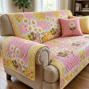 May include: A pink quilted sofa cover with a daisy floral design. The cover showcases white daisies with yellow centres and green stems, set against a pink backdrop. Yellow and white gingham trim borders the cover. A matching cushion with a daisy is also present.