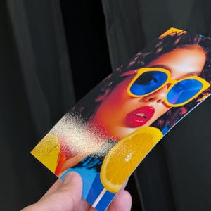 May include: A colourful print showing a woman wearing yellow sunglasses with blue lenses and red lipstick. A slice of lemon is also visible. The print is held against a black backdrop.