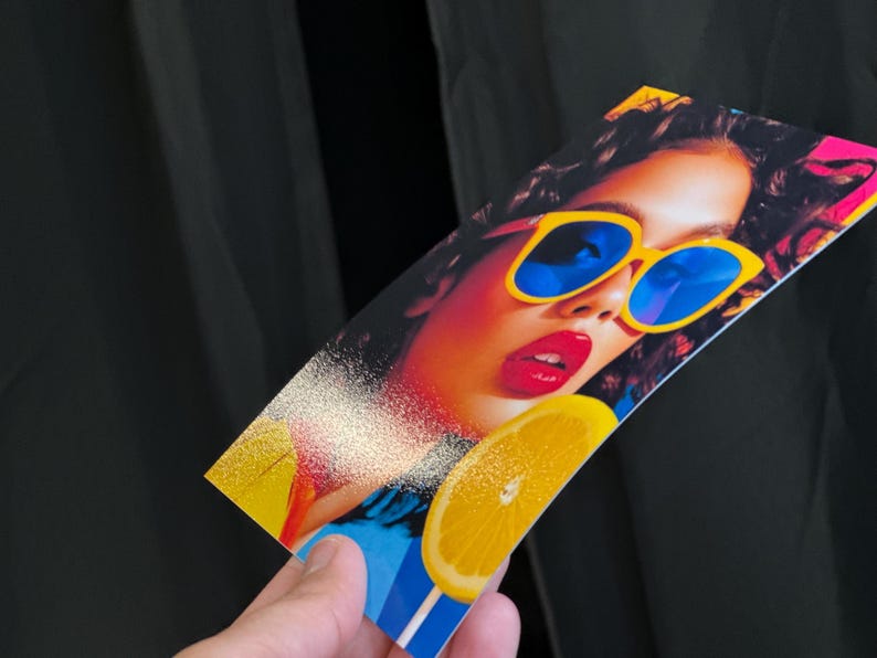 May include: A colourful print showing a woman wearing yellow sunglasses with blue lenses and red lipstick. A slice of lemon is also visible. The print is held against a black backdrop.