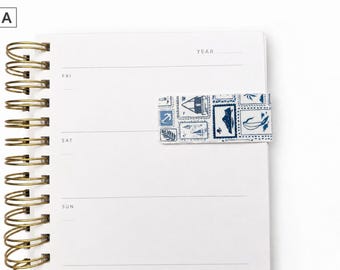 May include: A white spiral-bound notebook with a gold spiral and a blue and white patterned bookmark. The bookmark features a design of postage stamps. The notebook is open to a blank page with the days of the week printed on the left side.