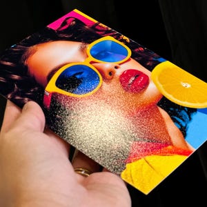 May include: A colourful print showing a woman with yellow sunglasses with blue lenses, red lipstick, and a slice of orange. The image has a glittery effect and is held against a black background.
