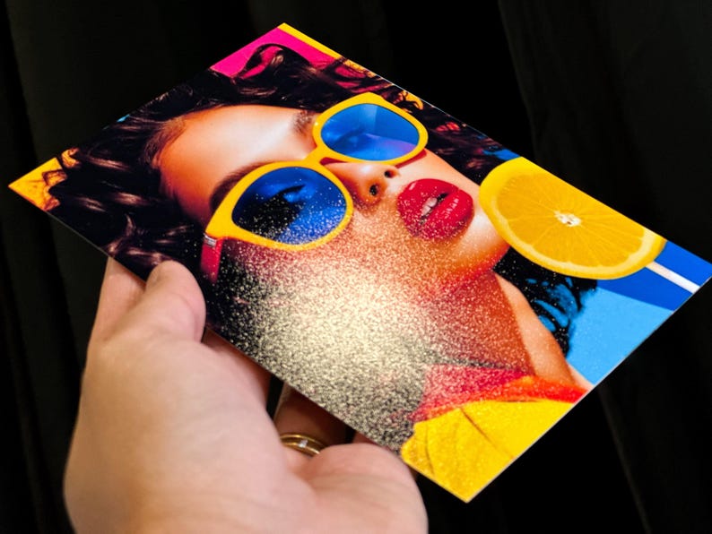 May include: A colourful print showing a woman with yellow sunglasses with blue lenses, red lipstick, and a slice of orange. The image has a glittery effect and is held against a black background.
