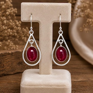 May include: A pair of silver drop earrings featuring a polished oval-shaped deep red gemstone suspended within a teardrop-shaped silver frame. The earrings hang from simple silver hooks and showcase a smooth, reflective finish on the stones.