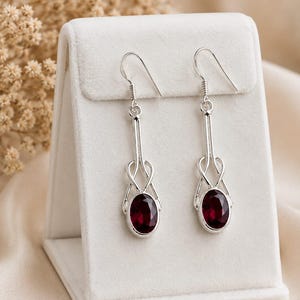 Peut inclure: Une paire de boucles d'oreilles pendantes en argent avec une monture en métal poli au design entrelacé. Chaque boucle est ornée d'une pierre précieuse rouge foncé de taille ovale à la base et suspendue par un crochet en argent.