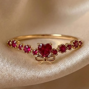 Puede incluir: Un anillo dorado con una piedra central en forma de corazón de color rubí y piedras rojas más pequeñas. El anillo presenta un diseño de lazo debajo del corazón y está sobre una tela color crema.