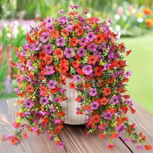 May include: A white ceramic pot overflowing with vibrant, cascading artificial flowers. The arrangement features a mix of red and purple blossoms with green foliage, creating a colorful display. The pot sits on a wooden surface.