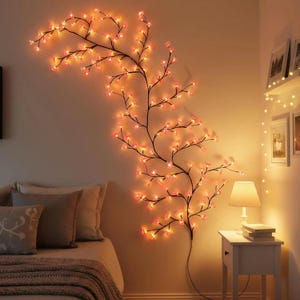 May include: A decorative wall-mounted light fixture shaped like a flowering branch. The branch is dark brown with pink and orange illuminated flowers. The fixture is mounted on a beige wall, with a bed and nightstand in the foreground.