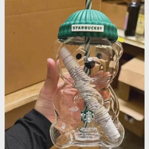 Peut inclure: Un gobelet en plastique transparent en forme d'ours avec un couvercle vert côtelé style bonnet portant le logo Starbucks. Une paille rayée verte et blanche est insérée. À l'intérieur se trouve un objet cylindrique blanc avec du texte imprimé.
