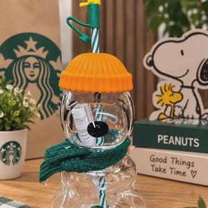 Peut inclure: Un gobelet en verre transparent Starbucks x Peanuts en forme de Snoopy, doté d'un couvercle orange vif en forme de bonnet côtelé, d'une écharpe en tricot vert autour du cou et d'une paille rayée vert et blanc surmontée d'une figurine jaune de Woodstock.