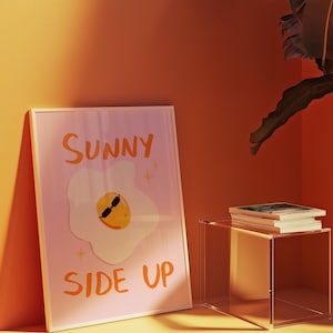 Peut inclure: Une affiche encadrée avec les mots "SUNNY SIDE UP" en lettres orange. L'œuvre d'art représente un œuf au plat de dessin animé portant des lunettes de soleil. Le fond est rose clair. L'affiche est affichée sur un mur orange.