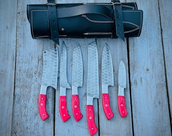 Pode incluir: Um conjunto de sete facas de cozinha em aço inoxidável com cabos de madeira vermelha, apresentado com um estojo de couro preto. As lâminas têm uma textura martelada. As facas estão dispostas sobre uma superfície de madeira envelhecida.