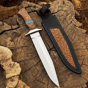 May include: A hunting knife with a polished steel blade and a wooden handle with turquoise accents. The knife is accompanied by a leather sheath with a reptile-skin pattern and teal stitching. The knife and sheath are on a textured wooden surface.