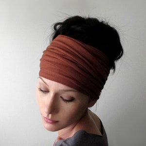 May include: A wide, rust-colored headband wraps around the head, contrasting with dark hair. The fabric appears soft and textured. The person is wearing a gray top and looking down, creating a serene and focused image.