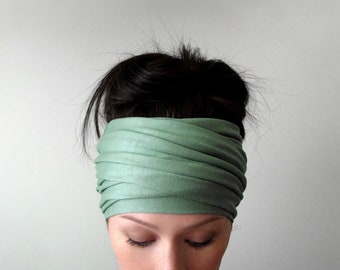 May include: A wide, sage green fabric headband featuring a series of horizontal, overlapping pleats that wrap around the head. The material appears soft and flexible, creating a layered, ruched texture across the entire width of the accessory.
