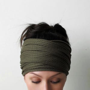 May include: An olive green headband featuring a textured waffle-knit fabric. The accessory is designed with a wide, gathered band that wraps around the head, creating a layered, crisscross appearance with multiple horizontal folds across the front.