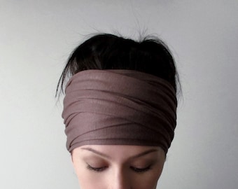 May include: A wide, solid brown fabric headband featuring a multi-layered, ruched, and wrapped design that covers the forehead and crown. The material appears soft and matte, with overlapping horizontal folds creating a textured, gathered appearance.