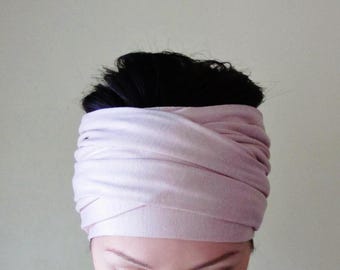 May include: A wide, pale pink fabric headband featuring a layered, wrapped design that covers the forehead and wraps around the head. The soft, solid-colored material is gathered in multiple horizontal folds, creating a textured appearance.