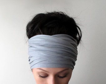 May include: A light gray headband featuring a wide, ruched fabric design that wraps around the head. The accessory is constructed from a soft, flexible material with multiple horizontal folds and a crisscross pattern across the front section.