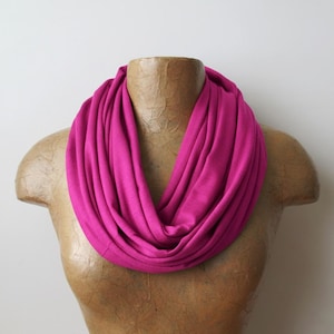 Op de afbeelding: Een fel magenta infinity sjaal gedrapeerd op een mannequin. De sjaal is gemaakt van een zachte, vloeiende stof, waardoor een volle, cirkelvormige vorm ontstaat. De heldere kleur van de sjaal is het middelpunt.