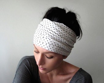 May include: A wide, stretchy fabric headband featuring a white base color with a repeating pattern of small, evenly spaced black polka dots. The headband is worn wrapped around the head, creating a gathered, layered appearance across the forehead.