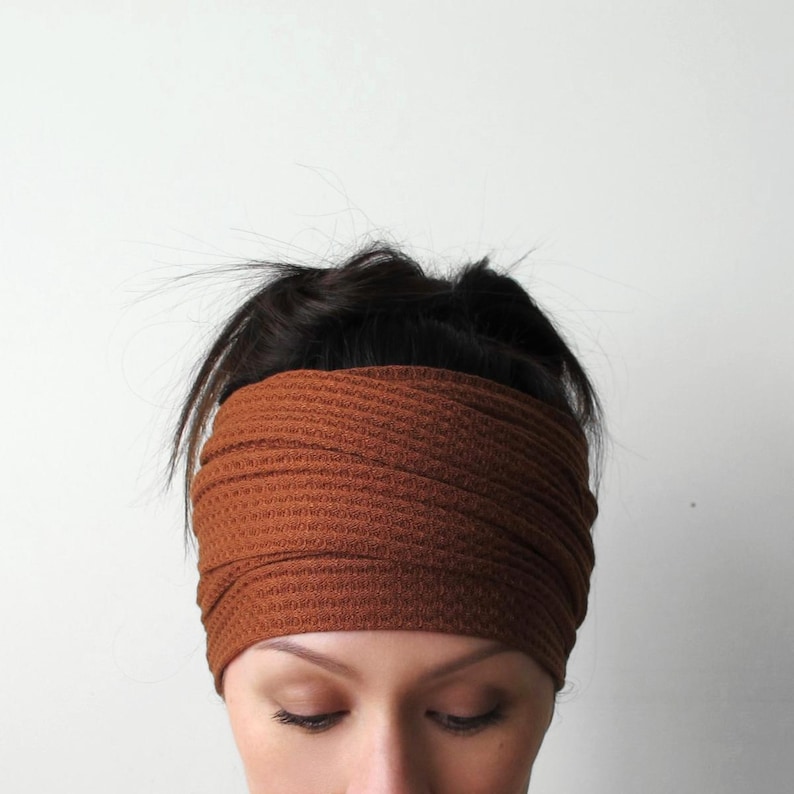 May include: A wide, rust-coloured headband featuring a textured waffle-knit fabric pattern. The accessory wraps around the head, covering the forehead and ears with a consistent, repeating grid-like weave throughout the entire surface of the material.