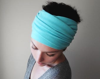 May include: A wide, solid light turquoise headband made of soft, stretchy fabric. The accessory features multiple horizontal folds and gathers, creating a ruched texture that wraps around the head to secure hair in a simple, functional style.