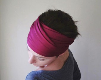 May include: A wide, deep magenta headband featuring a gathered, twisted fabric design that wraps around the head. The accessory is made of a smooth, solid-colored material with visible folds and pleats that create a layered, textured appearance.