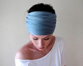 May include: A wide, dusty blue fabric headband featuring a gathered, multi-layered design that wraps around the head. The soft, matte material is arranged in horizontal folds, creating a textured appearance across the front and sides of the accessory.