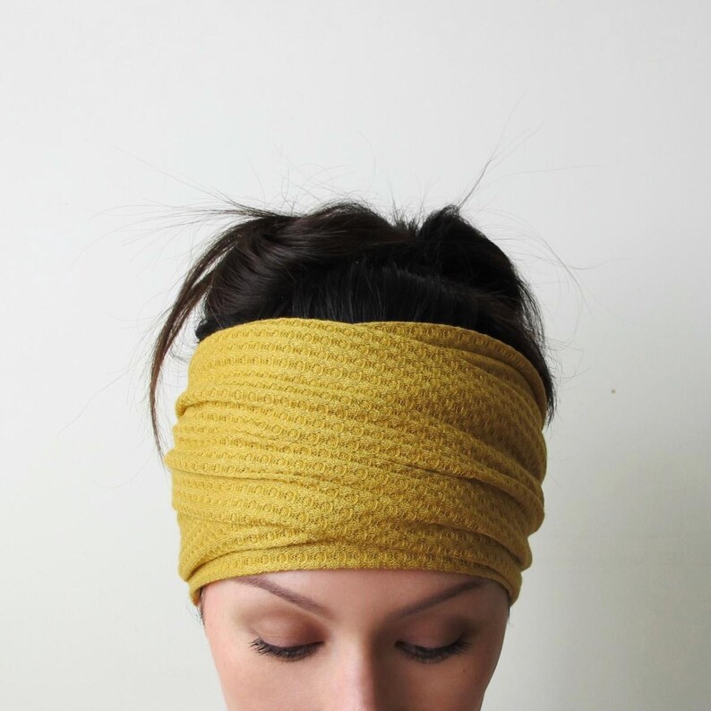 May include: A mustard yellow headband with a textured waffle-knit pattern. The fabric is gathered in horizontal folds, creating a layered appearance that wraps around the head. The material has a consistent, repeating grid-like structure throughout.