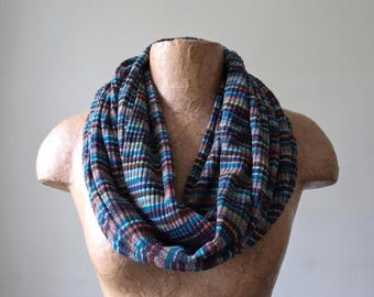 May include: A ribbed infinity scarf featuring a multi-colored striped pattern in shades of dark blue, teal, brown, and orange. The fabric has a textured, vertical knit construction that wraps around the neck in a continuous loop design.